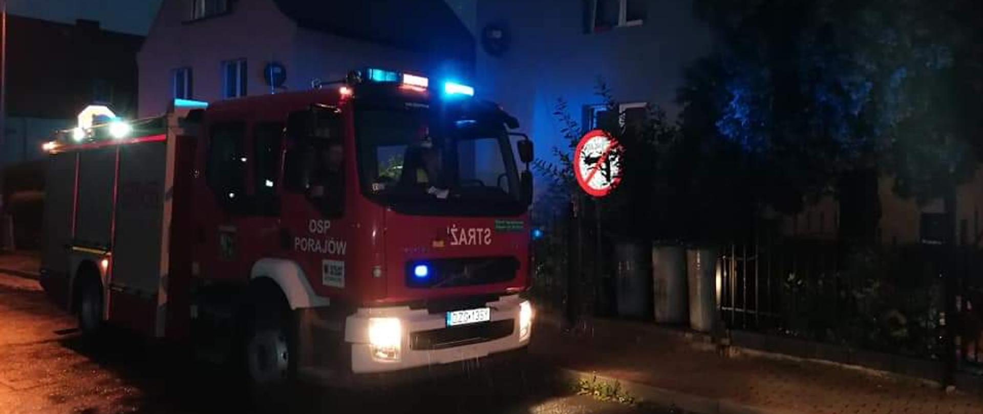 Pora nocna. Samochód ratowniczo gaśniczy z włączonymi światłami błyskowym. Na drzwiach samochodu napis OSP Porajów.