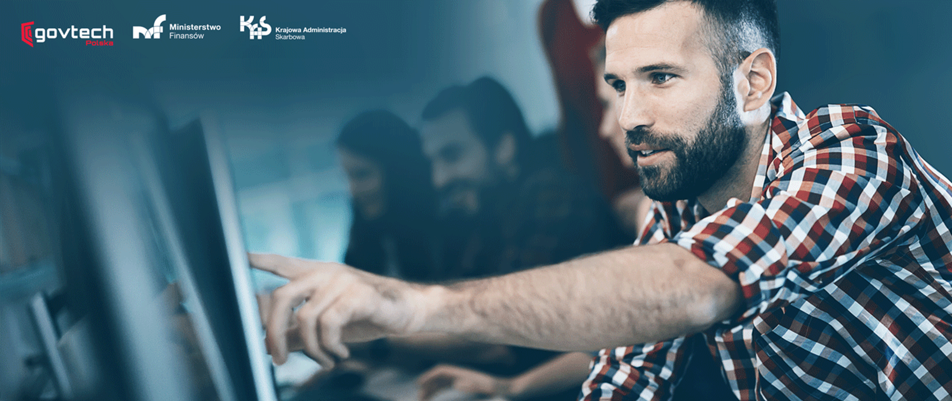 Mężczyzna wskazuje palcem na ekran komputera, za nim w tle inne osoby