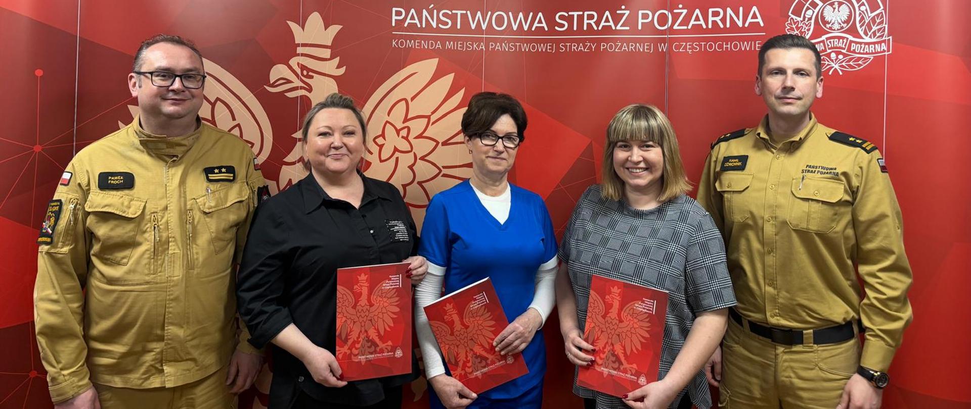 Na zdjęciu na tle czerwonej ścianki z napisem Państwowe Straż Pożarna ustawione trzy kobiety oraz dwóch mężczyzn w umundurowaniu służbowym. Kobiety w rękach trzymają czerwone teczki z białym orłem.