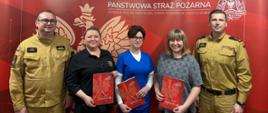 Na zdjęciu na tle czerwonej ścianki z napisem Państwowe Straż Pożarna ustawione trzy kobiety oraz dwóch mężczyzn w umundurowaniu służbowym. Kobiety w rękach trzymają czerwone teczki z białym orłem.