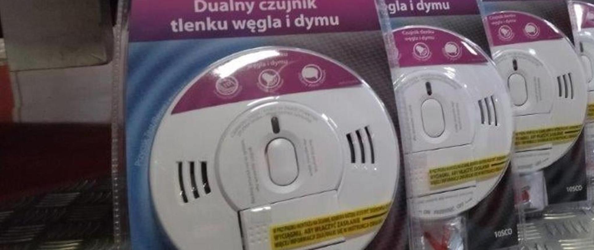 Sześć dualnych czujników tlenku węgla i dymu w plastykowych, przezroczystych opakowaniach ustawionych jest jedna za drugą na srebrnym podłożu.