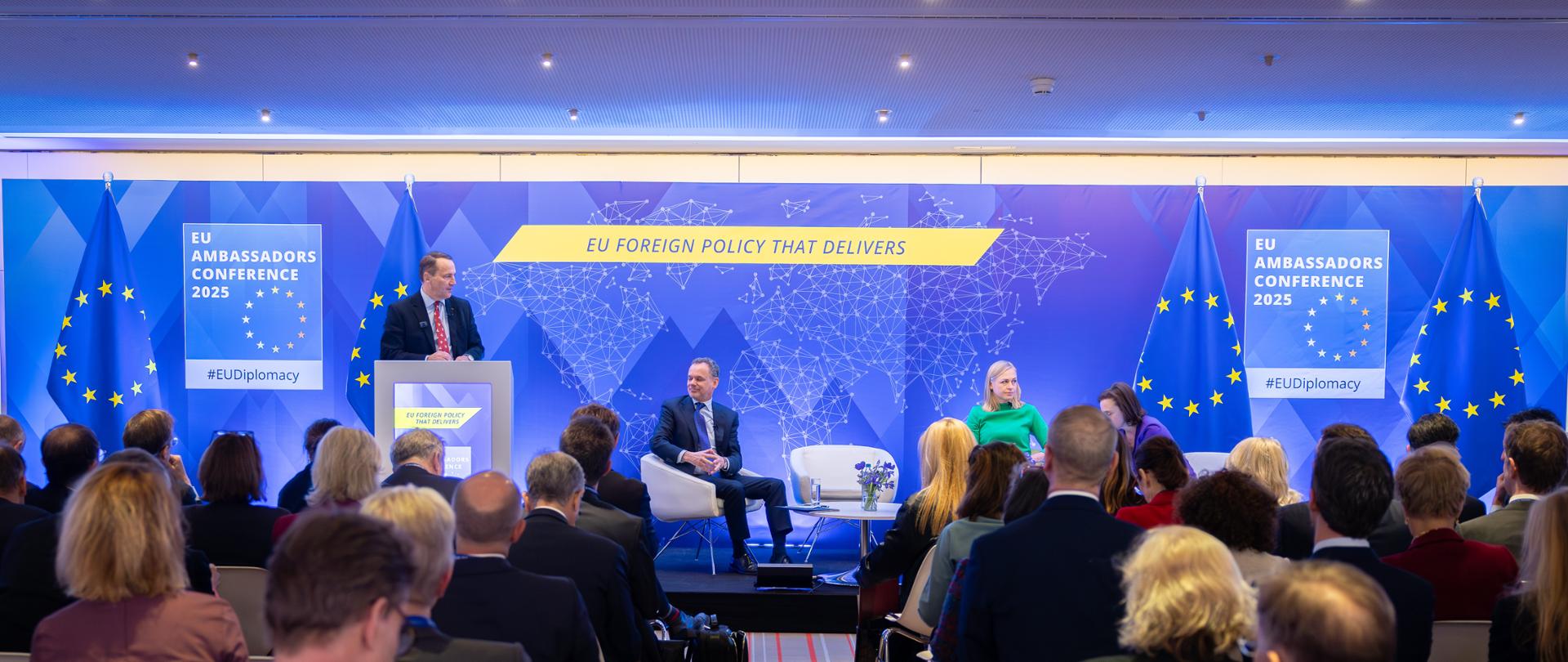 Minister Radosław Sikorski wziął udział w Konferencji Ambasadorów Unii Europejskiej. Na zdjęciu trzy osoby siedzą w fotelach obrotowych. W tle niebieska ścianka. Przy mównicy stoi minister Radosław Sikorski w garniturze.