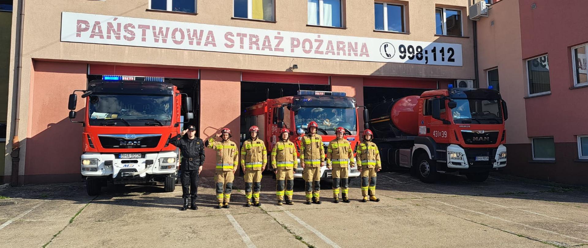 Sześciu strażaków w ubraniach specjalnych koloru piaskowego i hełmach na głowach koloru czerwonego oraz jeden w ubraniu dowódczo-sztabowym koloru czarnego stoi na baczność przed trzema samochodami pożarniczymi, które częściowo wyjechały z budynku. Samochody mają włączoną sygnalizację świetlną koloru niebeiskiego.