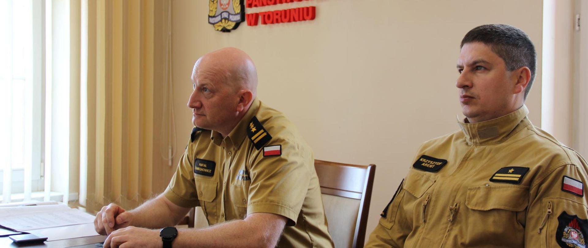 Przy stole konferencyjnym siedzi dwóch oficerów PSP w mundurach służbowych - eden notuje. Za nimi na ścinie napis z nazwą instytucji i logo.