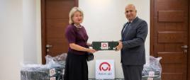 Dwie osoby stoją w pomieszczeniu i wspólnie trzymają zielone pudełko oznaczone logo Polish Aid. Za osobami znajdują się ułożone pudełka oraz duże elementy wyposażenia osłonięte przezroczystą folią. Na środku widoczna jest tablica z logo Polish Aid.