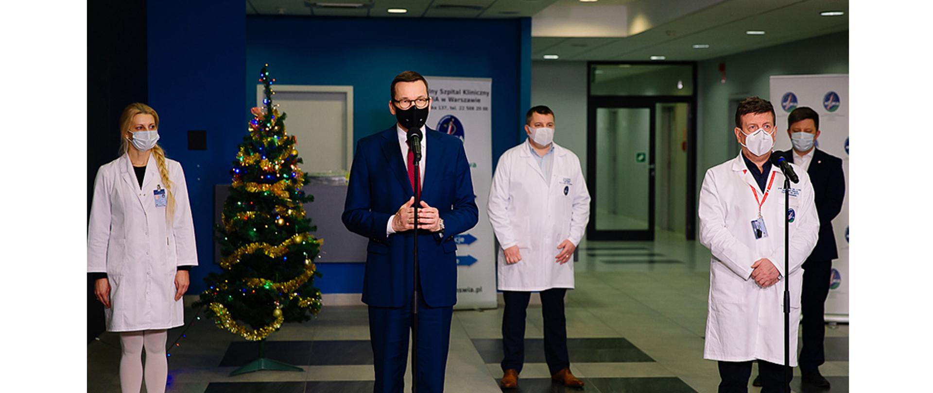 Na zdjęciu widać stojącego przed mikrofonem i przemawiającego premiera Mateusza Morawieckiego. Obok stoi przy mikrofonie dyrektor szpitala MSWiA w Warszawie prof. Waldemar Wierzba. W tle widać dwie osoby z personelu medycznego.
