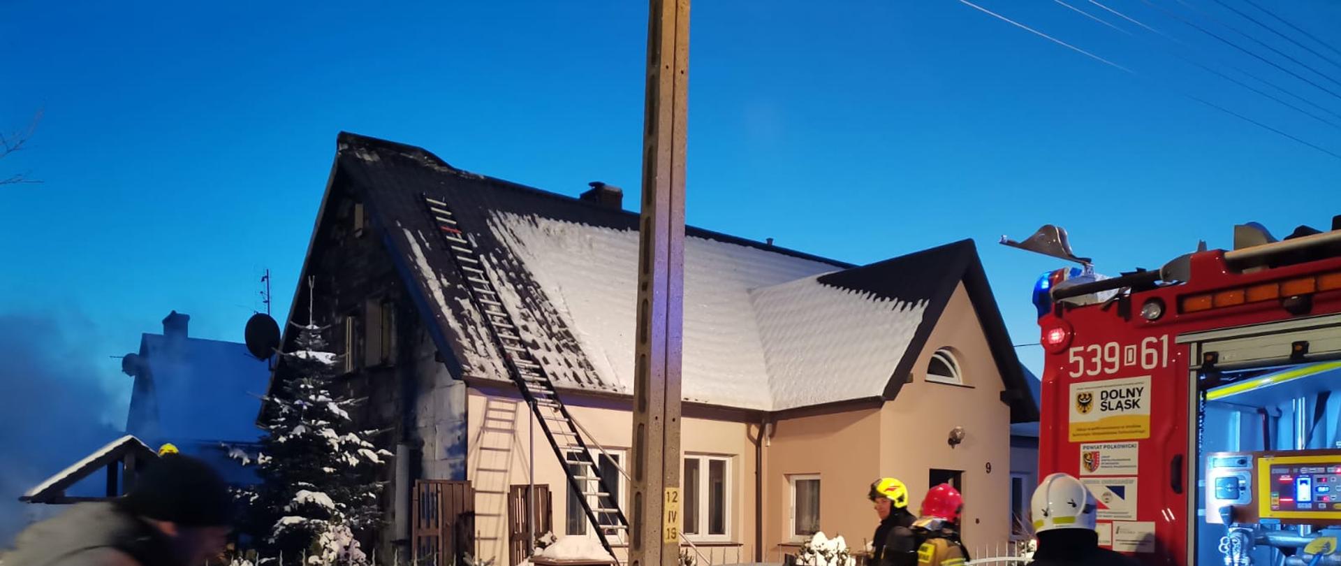 Zdjęcie przedstawia częściowo nadpalony dom jednorodzinny wraz ze strojącym pojazdem pożarniczym z rozwiniętymi wężami gaśniczymi. Przy aucie jest 3 strażaków. 