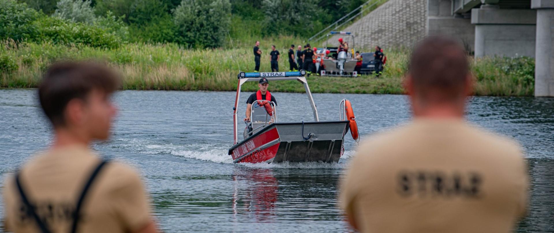 Na pierwszym planie widzimy łódź ratowniczą z OSP Łętowice, która wpływa na nurt Dunajca w kierunku brzegu, gdzie obserwatorami są uczestnicy ćwiczeń – ubrani w charakterystyczne koszulki z napisem „STRAŻ”. W tle, na brzegu rzeki, trwają przygotowania kolejnych zastępów do wodowania sprzętu oraz obserwacja działań. Po prawej stronie kadru widoczny fragment mostu drogowego – kluczowy punkt orientacyjny rejonu ćwiczeń.