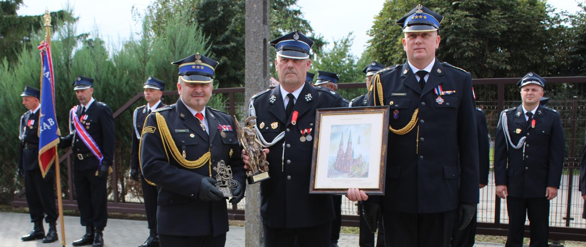 Uroczyste przekazanie pamiątkowych upominków z okazji 100-lecia OSP Obrąb. Na zdjęciu strażacy w galowych mundurach prezentują obraz oraz statuetkę, w tle stoją poczty sztandarowe i uczestnicy wydarzenia.