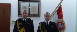 dwóch strażaków w ciemnych mundurach ze złotym sznurem , w białych koszulach z krawatem stoi obok siebie, w tle na ścianie sztandar oraz obraz 