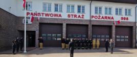 Zdjęcie przedstawia uroczystość przed budynkiem Państwowej Straży Pożarnej w Ostrowi Mazowieckiej, w dzień z częściowo zachmurzonym niebem. Na pierwszym planie, na brukowanym placu, stoi strażak w galowym mundurze, tyłem do widza, dowodząc grupą. Przed nim, w linii prostej, ustawionych jest około dziesięciu strażaków w ciemnych, bojowych mundurach z żółtymi elementami oraz w piaskowych mundurach polowych, niektórzy z nich salutują.
Budynek straży pożarnej jest biały z elementami ceglanymi i brązowym dachem. Nad trzema dużymi, brązowymi bramami garażowymi widnieje czerwony napis "PAŃSTWOWA STRAŻ POŻARNA" oraz numer alarmowy "998". Na fasadzie budynku, nad jedną z bram, wisi mała polska flaga. Z lewej strony budynku widoczny jest maszt flagowy, na którym również powiewa polska flaga. Niebo jest częściowo błękitne z widocznymi jasnymi chmurami.