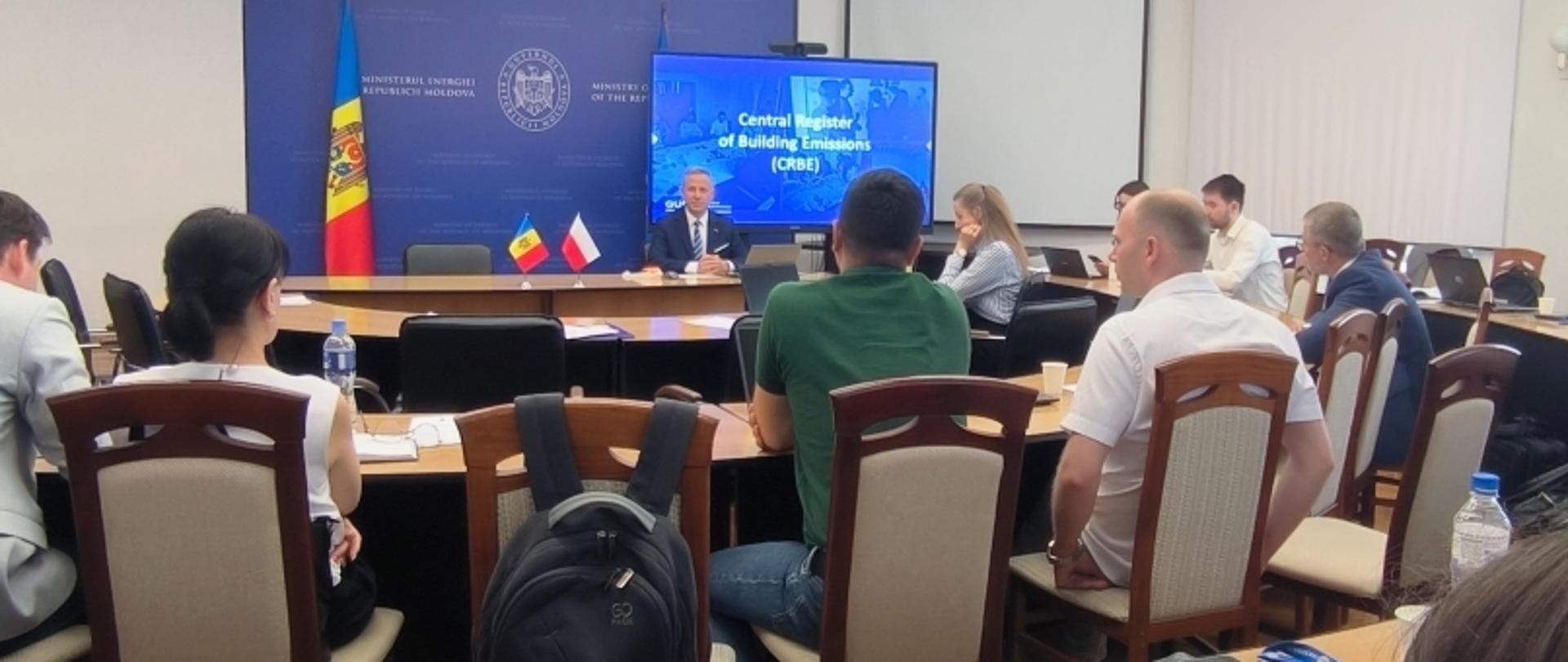 Sala konferencyjna z uczestnikami siedzącymi wokół dużego, zakrzywionego stołu. Na przodzie mężczyzna przemawia z mównicy, za którą znajdują się dwie flagi: Mołdawii i Polski. Na ekranie za nim widnieje napis 'Central Register of Building Emissions (CRBE)'. Pomieszczenie jest jasne, z białymi ścianami i naturalnym światłem wpadającym przez okna po prawej stronie.