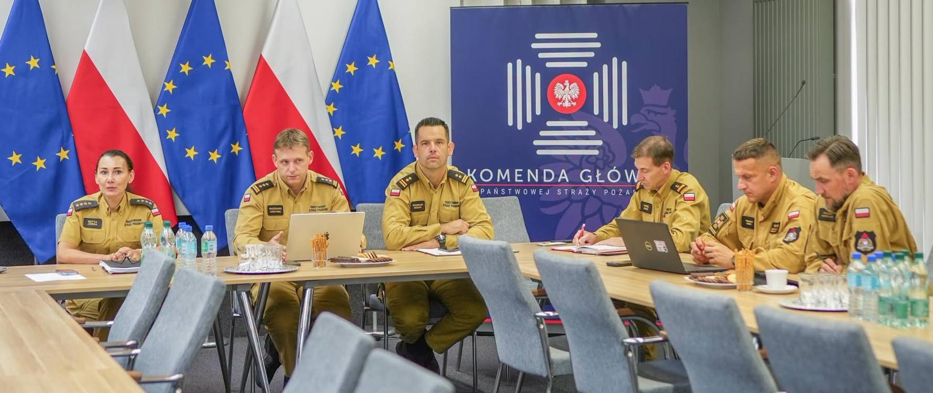 Widok ogólny sali konferencyjnej podczas spotkania w Komendzie Głównej Państwowej Straży Pożarnej. Przy stole siedzą funkcjonariusze PSP w umundurowaniu koszarowym. W tle widoczne są flagi Polski i Unii Europejskiej, godło państwowe oraz baner KG PSP.