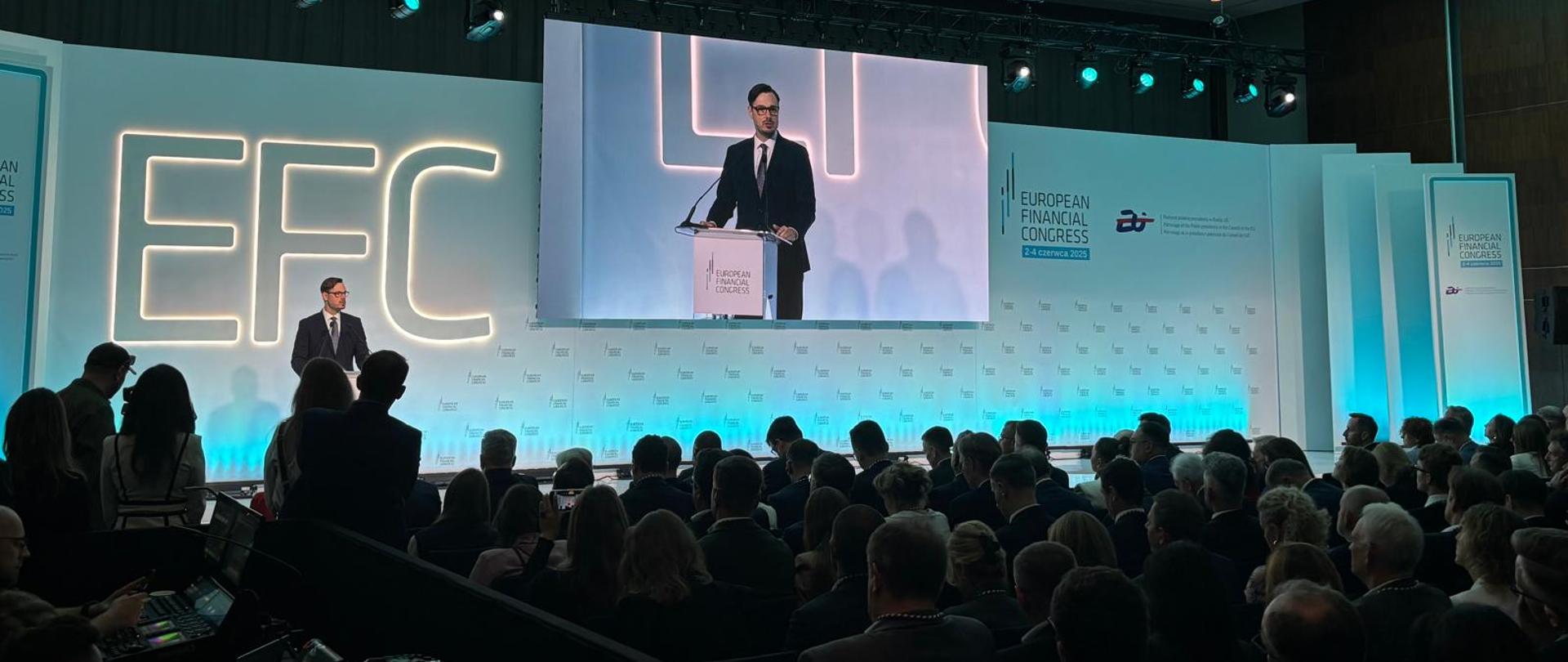 Minister Jakub Jaworowski stoi za mównicą. W tle ekran. Ujęcie zza słuchaczy. 