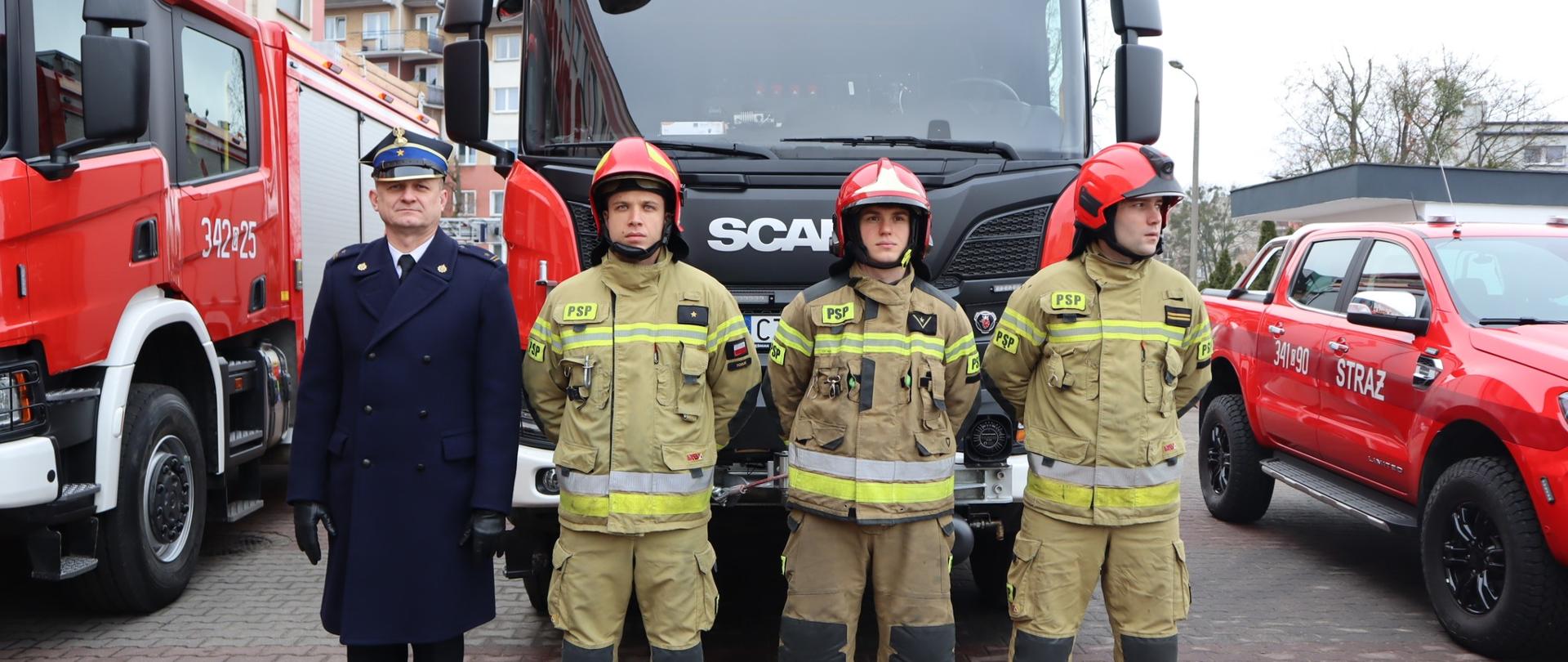 Dowódca ubrany w mundur wyjściowy i trzy-osobowa załoga wozu w ubraniu specjalnym. W tle samochód pożarniczy.