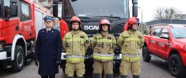 Dowódca ubrany w mundur wyjściowy i trzy-osobowa załoga wozu w ubraniu specjalnym. W tle samochód pożarniczy.