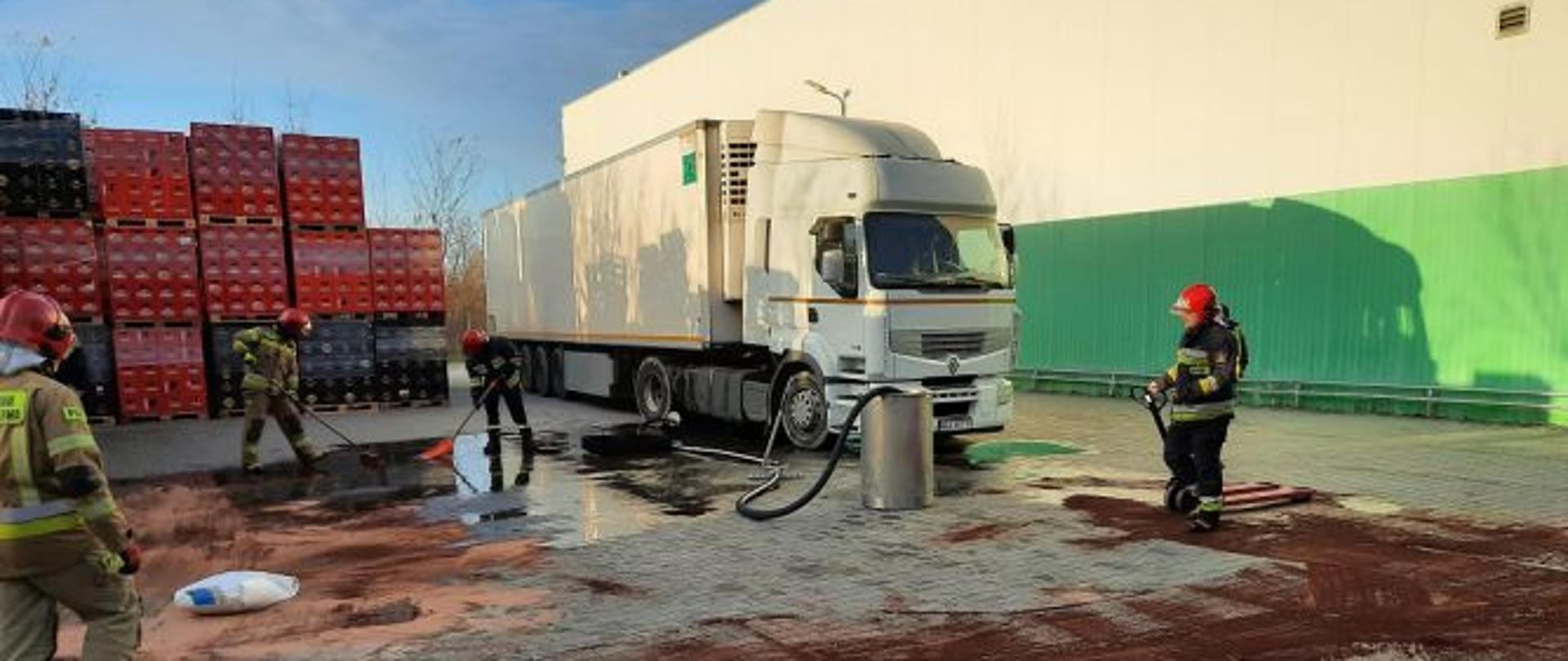 Ratownicy ze specjalistycznej grupy ratownictwa chemiczno-ekologicznego "SKARŻYSKO" podczas działań usuwania skutków wycieku paliwa z uszkodzonego zbiornika w samochodzie ciężarowym, do którego doszło na placu hurtowni Eurocash S.A w Skarżysku Kamiennej. Na placu wyłożonym kostką brukową, na którym doszło do wycieku stoi ciężarowy samochód ciężarowy z naczepą. Przy pojeździe ratownicy ustawili specjalistyczny sprzęt do przepompowania oleju napędowego z uszkodzonego zbiornika paliwa samochodu ciężarowego do zastępczego pojemnika. Za samochodem widać ustawioną piramidę z platikowych skrzynek transportowych w kolorze czarnym i czerwonym. W obrębie rozlewiska które powstało przy stojącym samochodzie ciężarowym czterech ratowników w czerwonych hełmach i ubraniach bojowych prowadzi działania ratownicze zabezpieczające wyciek i powstałe rozlewisko oleju napędowego.