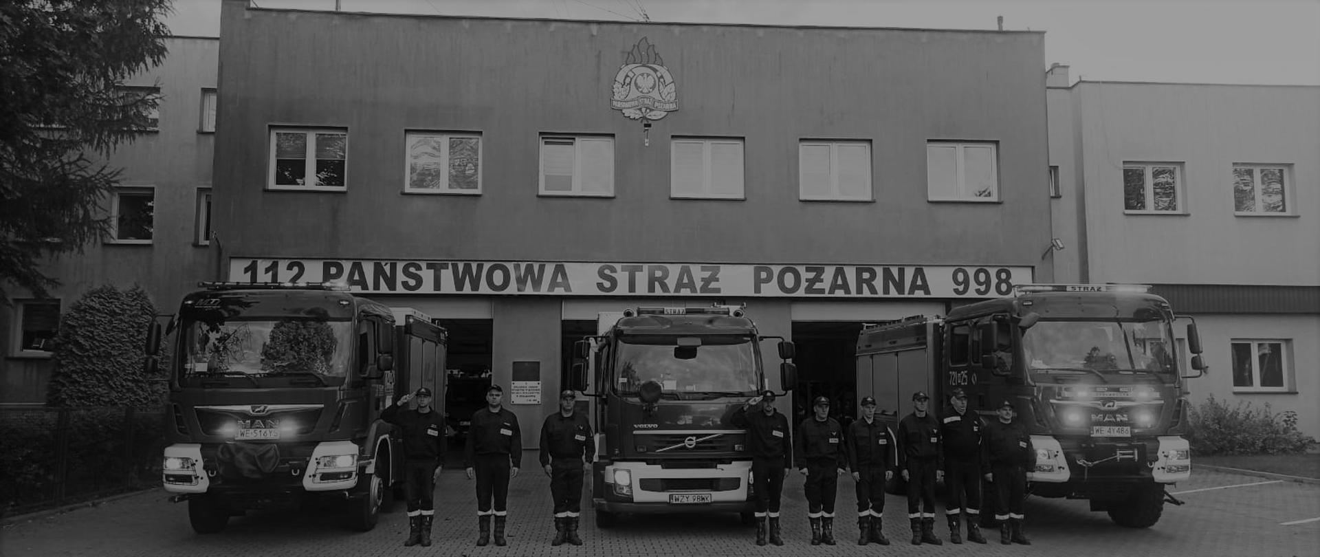 Zdjęcie wykonane przed Komendą Powiatową Państwowej Straży Pożarnej w Żyrardowie. Na zdjęciu widać strażaków ustawionych w szeregu przed jednostką. Strażacy ubrani w ubrania koszarowe. Za nimi ustawione są pojazdy ratowniczo – gaśnicze. Pojazdy mają włączone alarmowe sygnały świetlne i dźwiękowe. Strażacy oddają hołd zmarłemu koledze strażakowi.