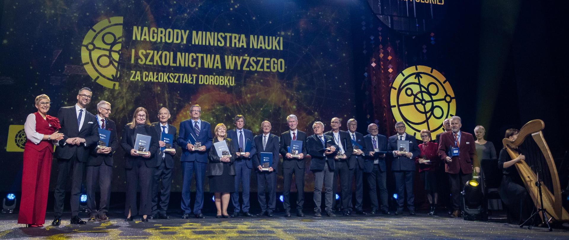 Na oświetlonej sali stoi w rzędzie duża grupa ludzi, po lewej minister Kulasek i wiceminister Mrówczyńska w czerwonej sukni, dalej rząd ludzi trzymających niebieskie teczki, za nimi na wielkim niebieskim ekranie żółty napis Nagrody Ministra Nauki i Szkolnictwa Wyższego.