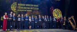 Na oświetlonej sali stoi w rzędzie duża grupa ludzi, po lewej minister Kulasek i wiceminister Mrówczyńska w czerwonej sukni, dalej rząd ludzi trzymających niebieskie teczki, za nimi na wielkim niebieskim ekranie żółty napis Nagrody Ministra Nauki i Szkolnictwa Wyższego.