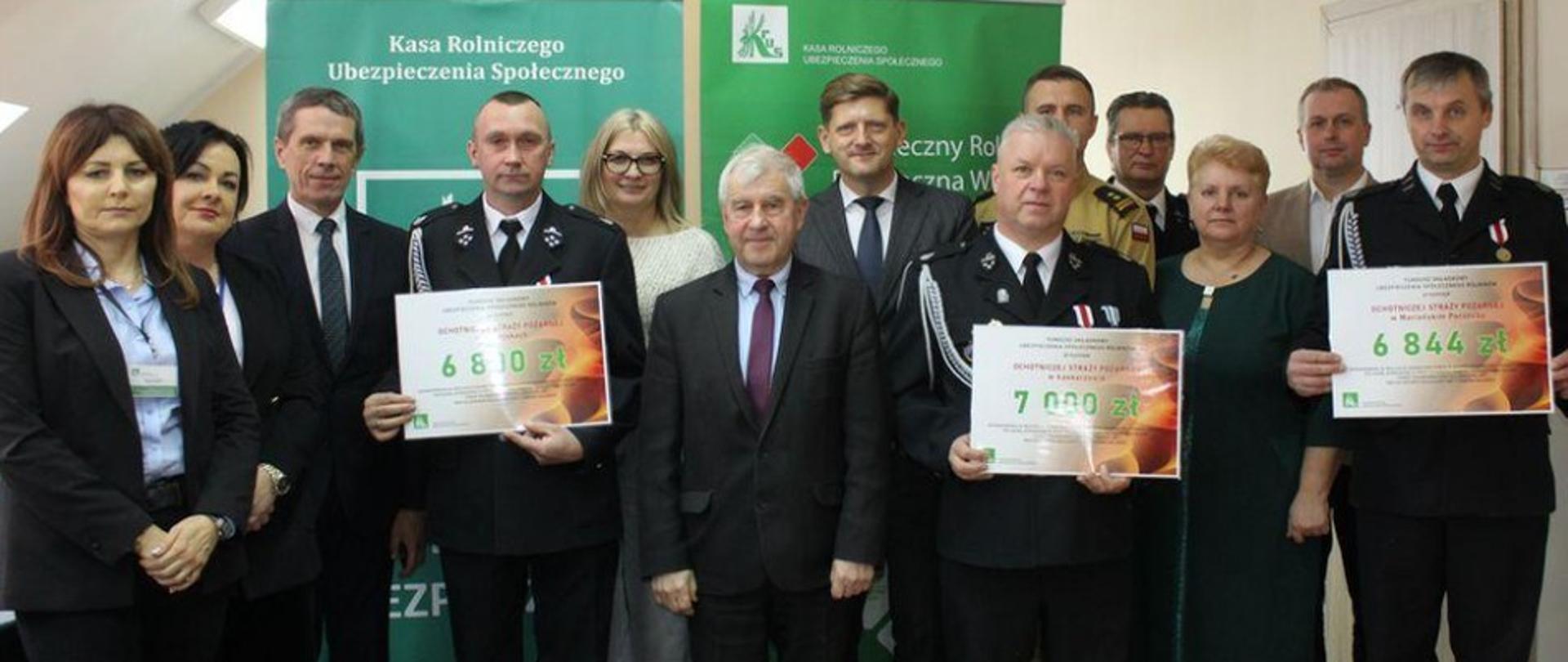 9 mężczyzn i 3 kobiety. Trzech mężczyzn z OSP Ryczyska, Mariańskie Porzecze i Łaskarzew trzymają w rękach vouchery z napisaną kwotą dofinansowania dla ich jednostek OSP.