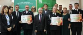 9 mężczyzn i 3 kobiety. Trzech mężczyzn z OSP Ryczyska, Mariańskie Porzecze i Łaskarzew trzymają w rękach vouchery z napisaną kwotą dofinansowania dla ich jednostek OSP.