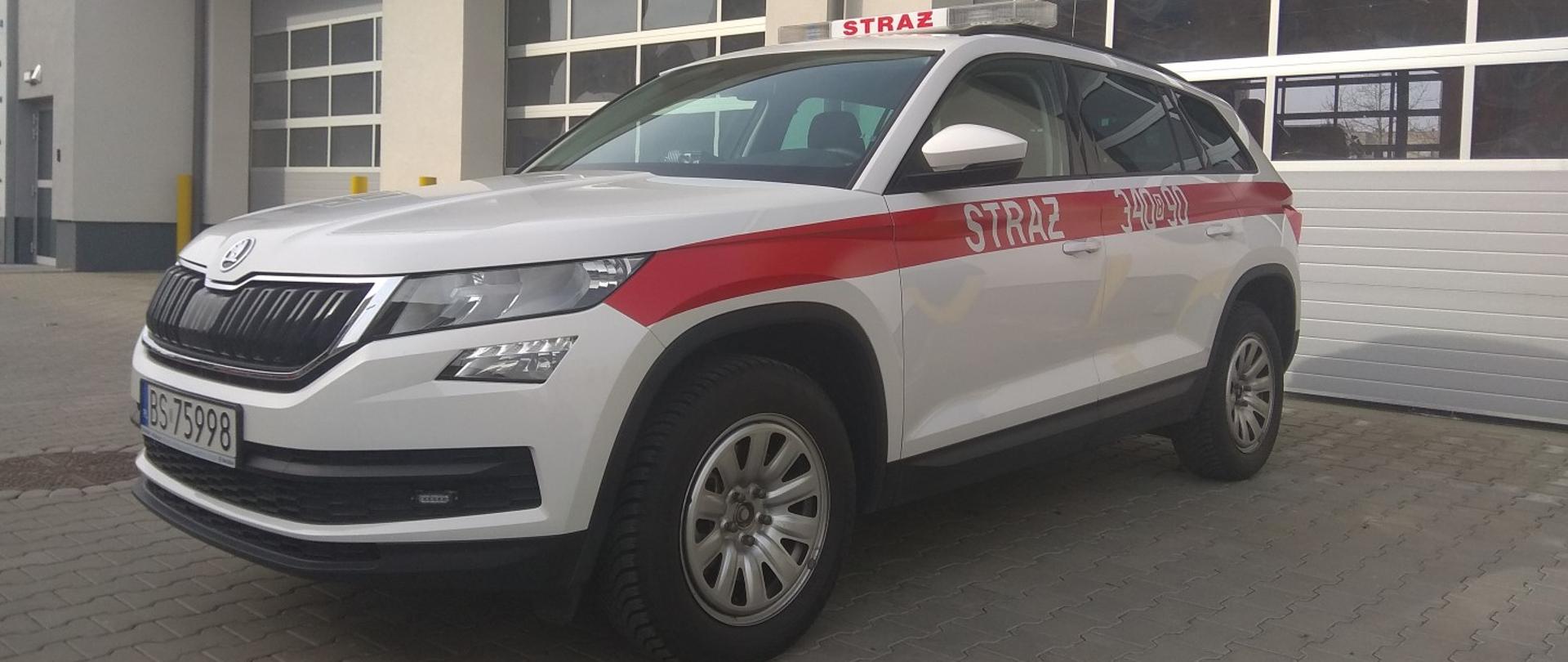 Na pierwszym planie samochód osobowy koloru białego z czerwonym paskiem pośrodku pojazdu. W tle zdjęcia garaże. 