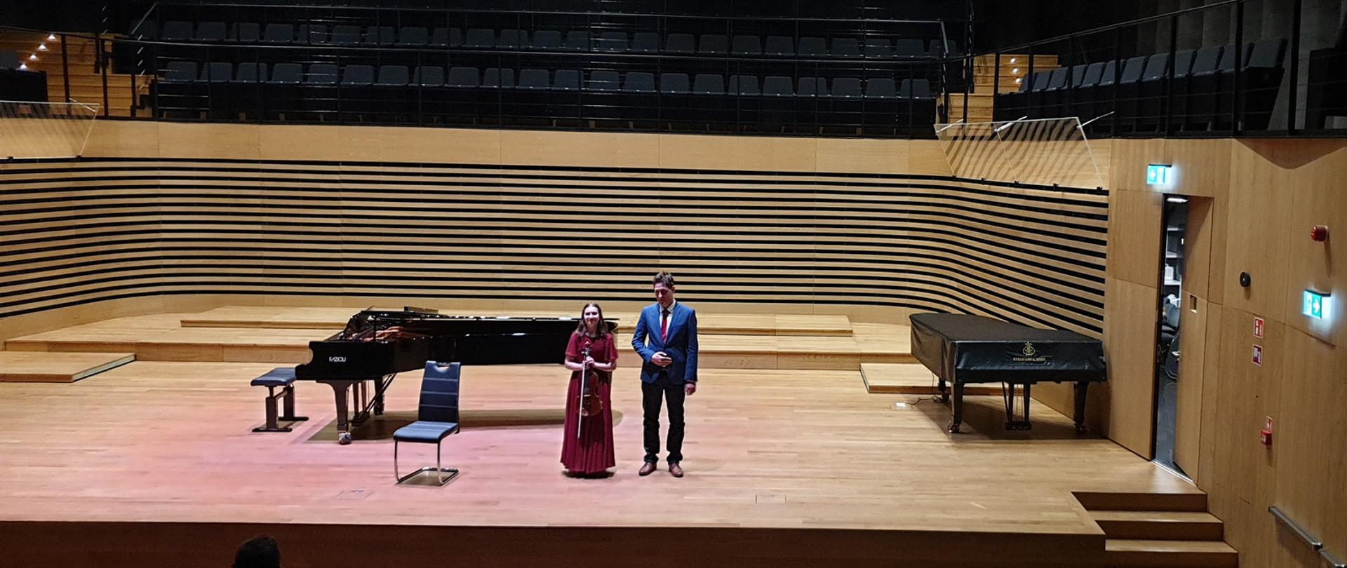 Dziewczyna stojąc trzyma w rękach przed sobą altówkę, obok stoi młody mężczyzna, za nimi stoi fortepian, po prawej stronie drugi fortepian.