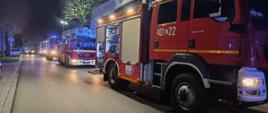Widok na ulicę, na której rozstawione są pojazdy strażaków. Jest noc. Auto stoją w szeregu, jedno za drugim. W szeregu widać auta gaśnicze i podnośnik.