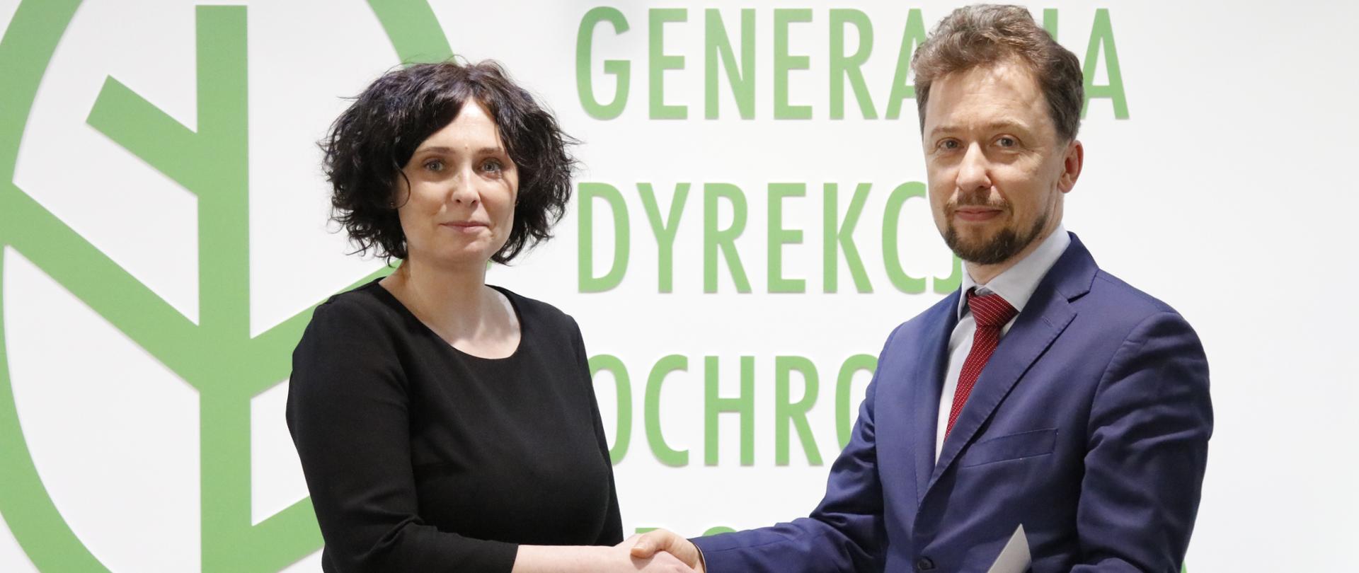 Na tle logotypu Generalnej Dyrekcji Ochrony Środowiska stoją kobieta i mężczyzna. Mężczyzna w granatowym garniturze wręcza kobiecie teczkę. Kobieta ubrana jest w czarną sukienkę.