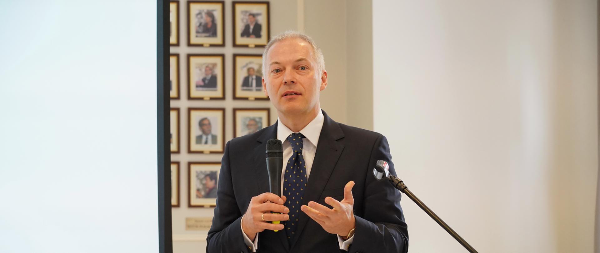  Wiceminister Jacek Żalek stoi przy mównicy i mówi do mikrofonu. W tle w małych ramkach zdjęcia bankowców.