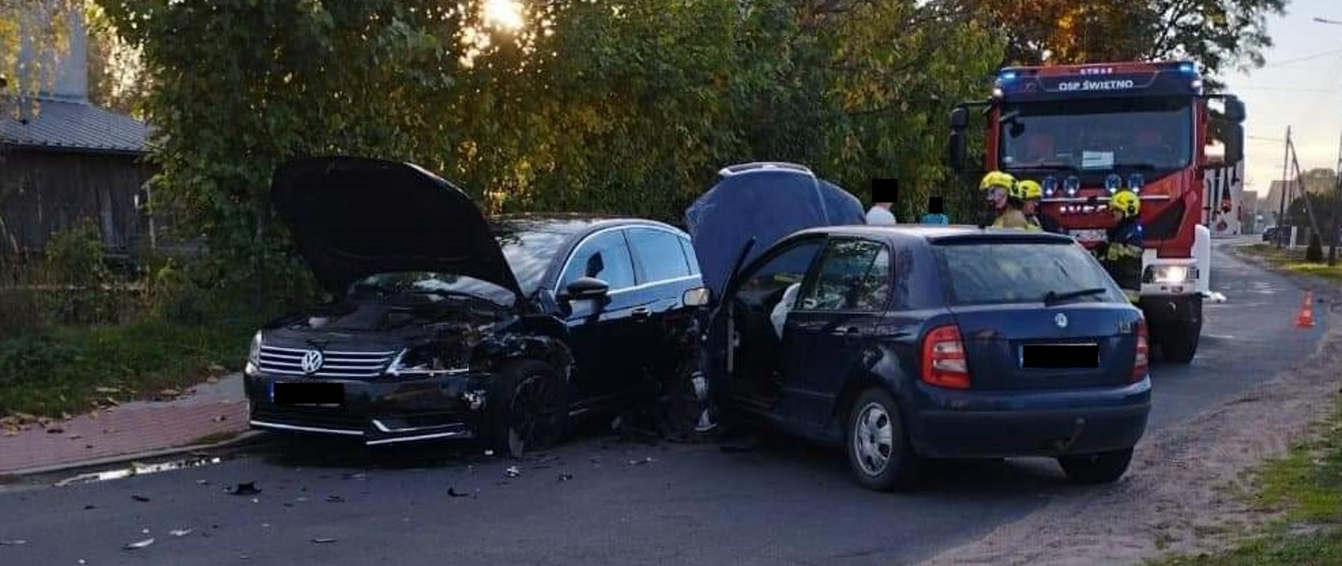 Zdjęcie przedstawia dwa samochody osobowe, rozbite, stojące na drodze. Za nimi znajduje się samochód strażacki.