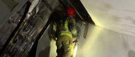 Na zdjęciu strażak stojący na drabinie, demontujący elementy obudowy konstrukcji dachu od wnętrza pomieszczenia.