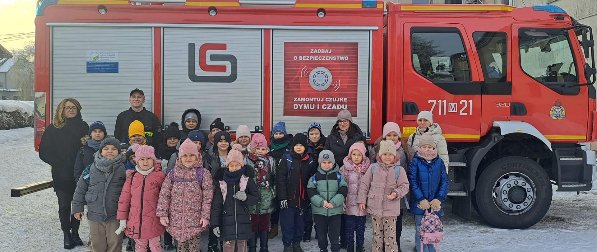Zdjęcie przedstawia grupę dzieci i dorosłych stojących na śniegu przed dużym, czerwonym wozem strażackim z napisem promującym montaż czujek dymu i czadu. Wszyscy są ubrani w ciepłą, zimową odzież, a w tle widoczne są ośnieżone budynki i drzewa