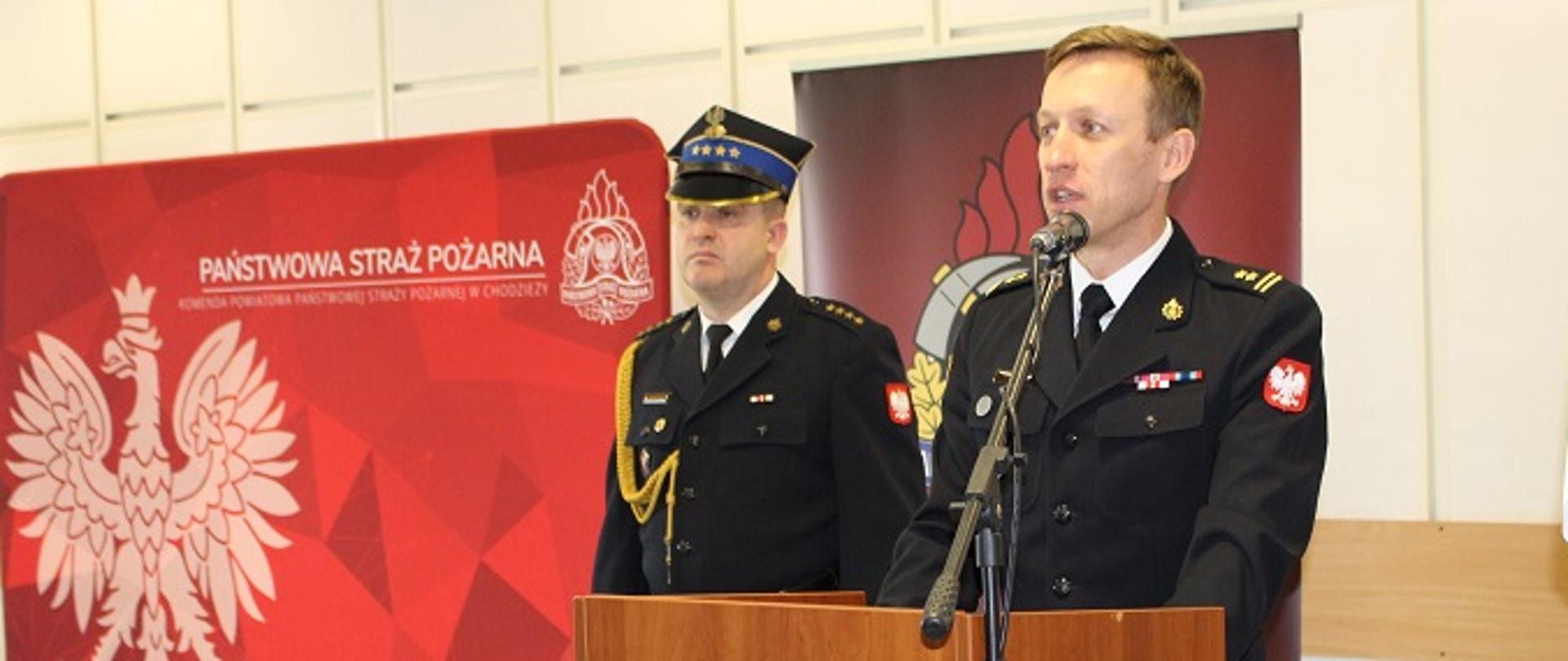 strażak stoi przy mównicy, za nim stoi inny strażak w rogatywce, w tle czerwony baner z orłem 