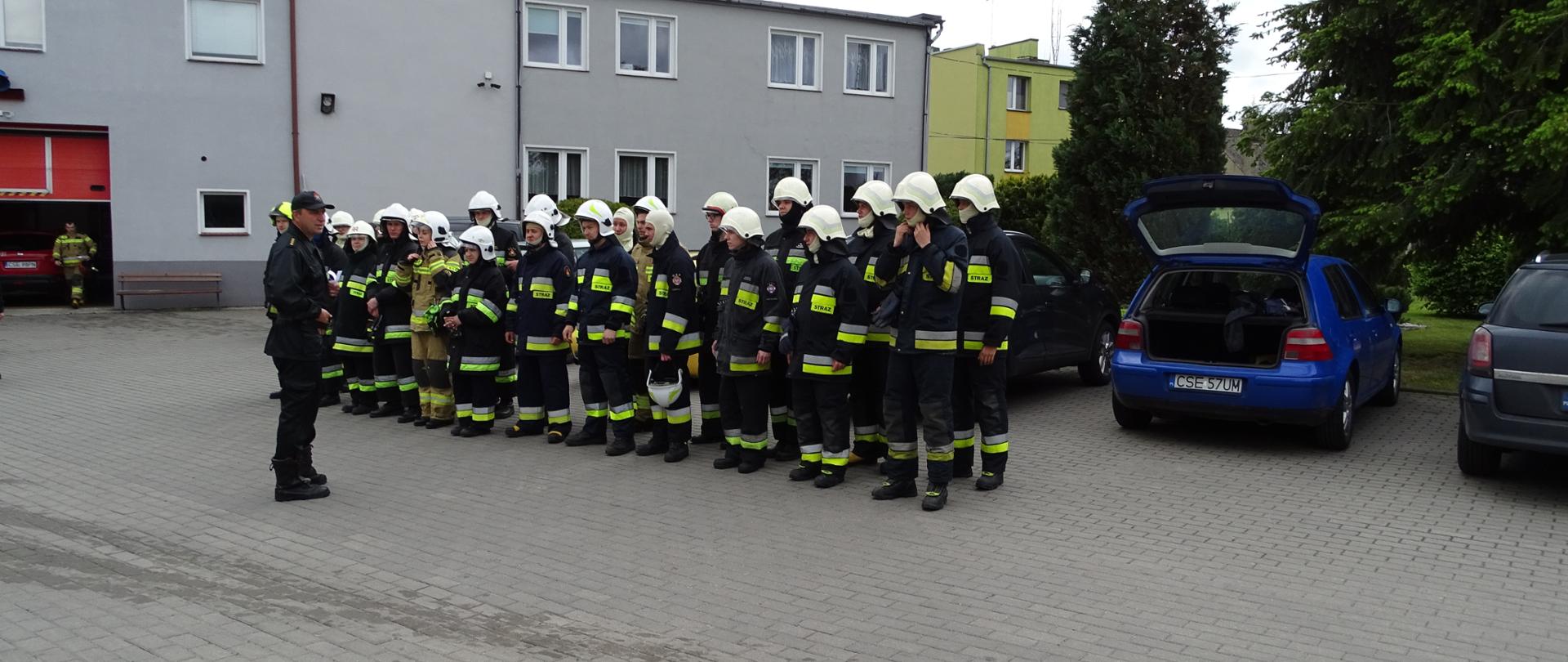 zdjęcie wykonane w dzień na placu komendy. Zdjęcie przedstawia strażaków osp w umundurowaniu bojowym podczas zbiórki przed egzaminem końcowym. Przed strażakami stoi zastępca dowódcy JRG w umundurowaniu dowódczo-sztabowym. W tle budynki komendy i oczko wodne. Po prawej stronie samochody prywatne.