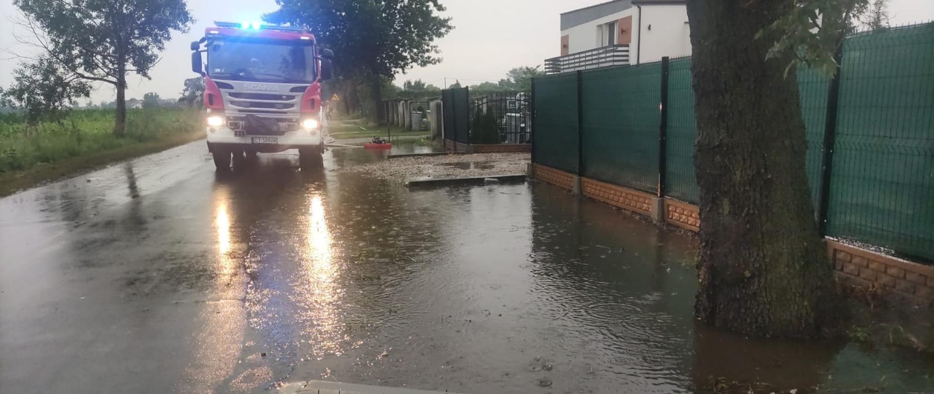 Zdjęcie przedstawia jezdnie drogi powiatowej na której znajduje się pojazd ratowniczo-gaśniczy Państwowej Straży Pożarnej z Radziejowa w trakcie zdarzenia zalanej posesji oraz jedni drogi powiatowej.