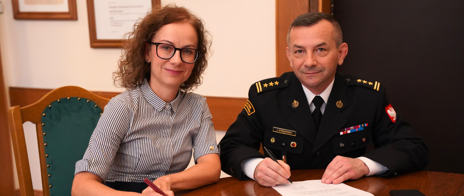 Pani Rektor i Pan Komendant, zajmując miejsca przy stole, podpisują porozumienie o współpracy, na dwóch egzemplarzach. 