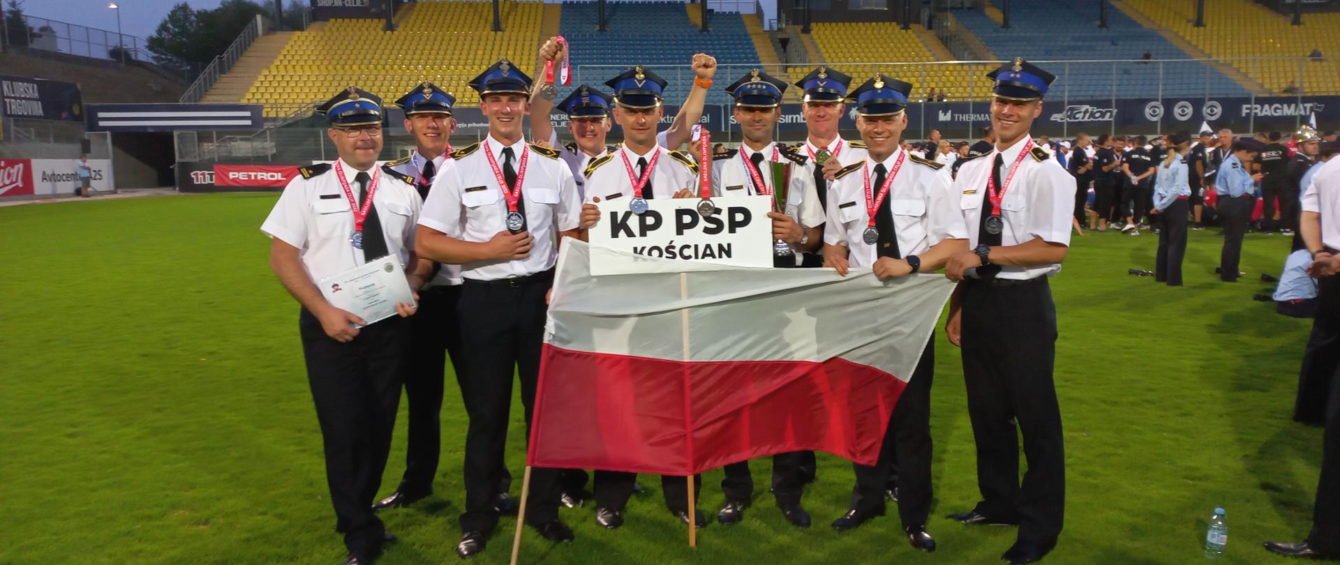 SUKCES KOŚCIAŃSKICH STRAŻAKÓW. Na zdjęciu widzimy strażaków na płycie boiska, ubrani w stroje służbowe, spodnie, biała koszula, czapka rogatywka, na szyi mają zawieszone srebrne medale. przed sobą trzymają tabliczkę z napisem KP PSP Kościan oraz biało-czerwoną flagę.