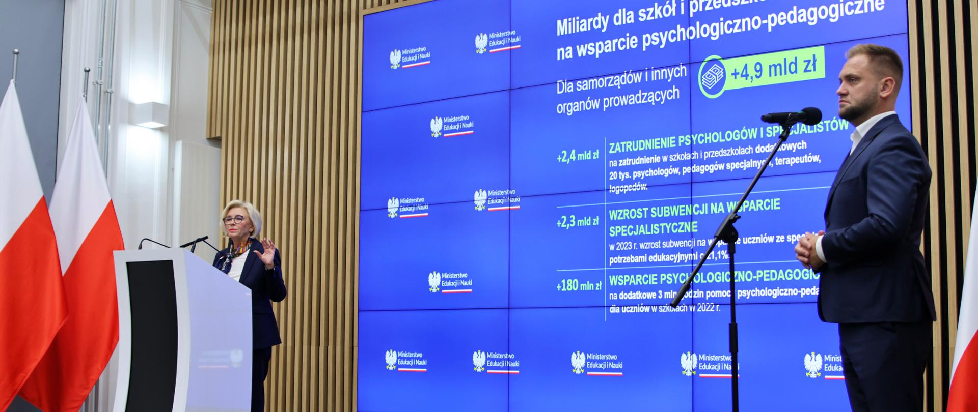 Widok z boku pod kątem na salę, wiceminister Machałek stoi za mównicą i mówi do dwóch mikrofonów, za nią wielki niebieski ekran z napisem Miliardy dla szkół i przedszkoli, z boku przed mikrofonem na stojaku stoi mężczyzna w garniturze.