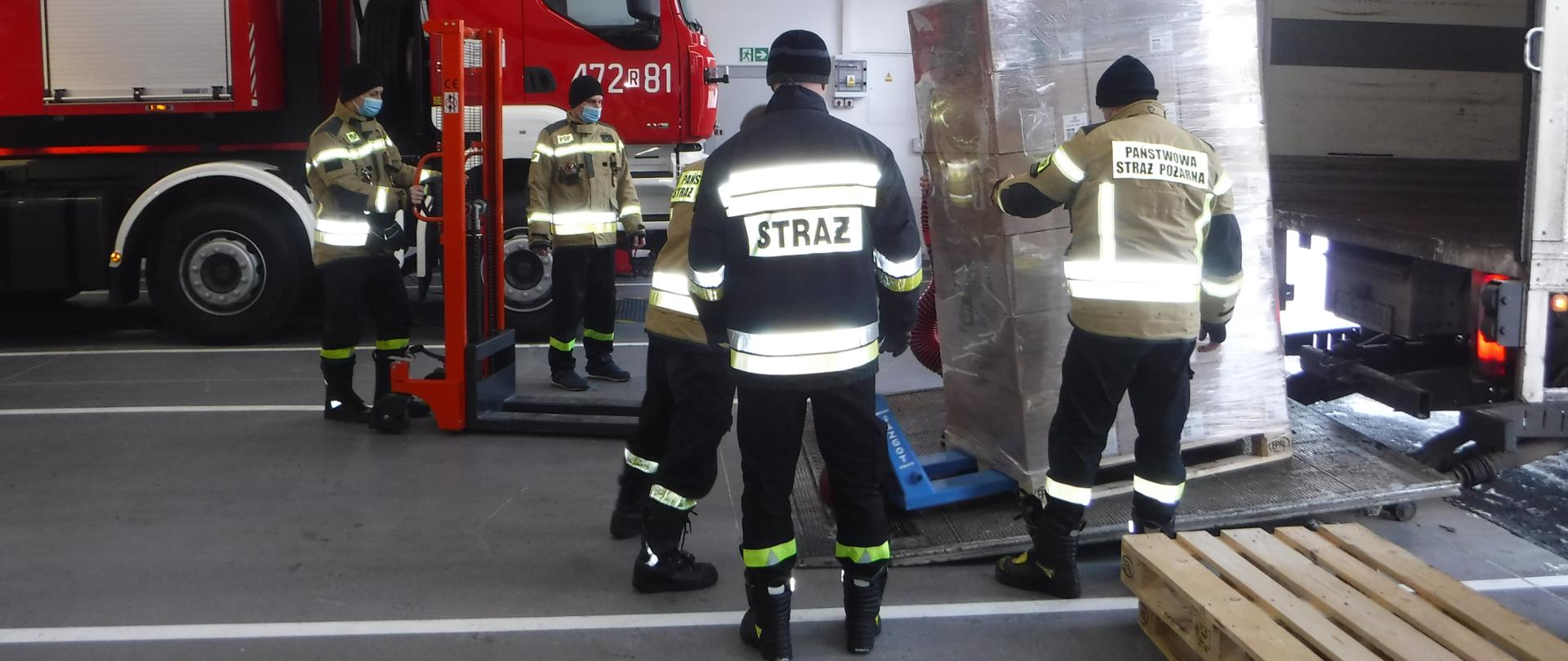 Na zdjęciu widzimy strażaków, którzy za pomocą wózków paletowych rozładowują samochód ciężarowy z ładunku maseczek jednorazowych.
