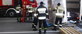 Na zdjęciu widzimy strażaków, którzy za pomocą wózków paletowych rozładowują samochód ciężarowy z ładunku maseczek jednorazowych.