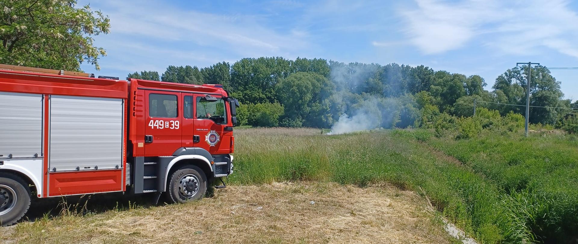Na zdjęciu widać czerwony samochód strażacki stojący na łące. W oddali widać dym unoszący się nad łąką. W tle widać drzewa, krzewy i linie energetyczne. Jest widno.