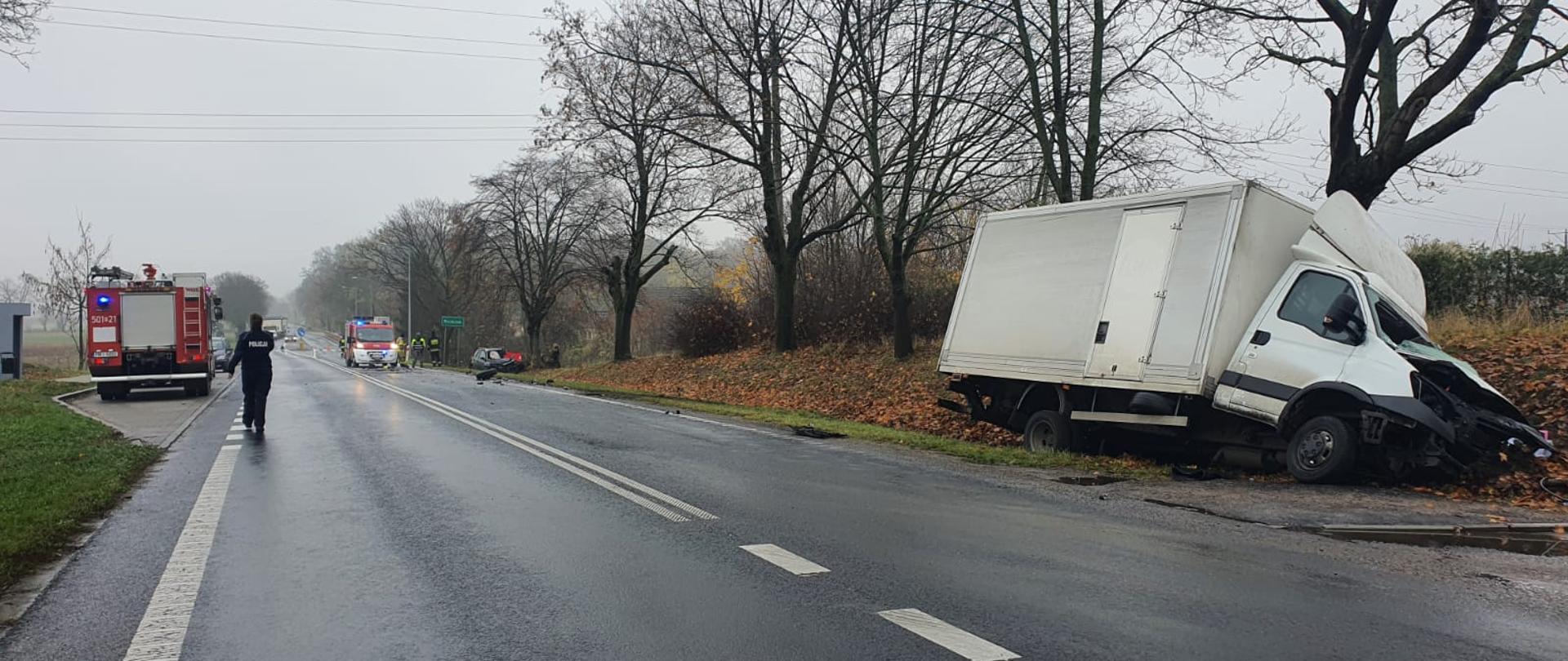 Na zdjęciu widać samochód dostawczy, który znajduje się w rowie. W oddali widać kolejny rozbity pojazd osobowy, policję i samochody strażackie.