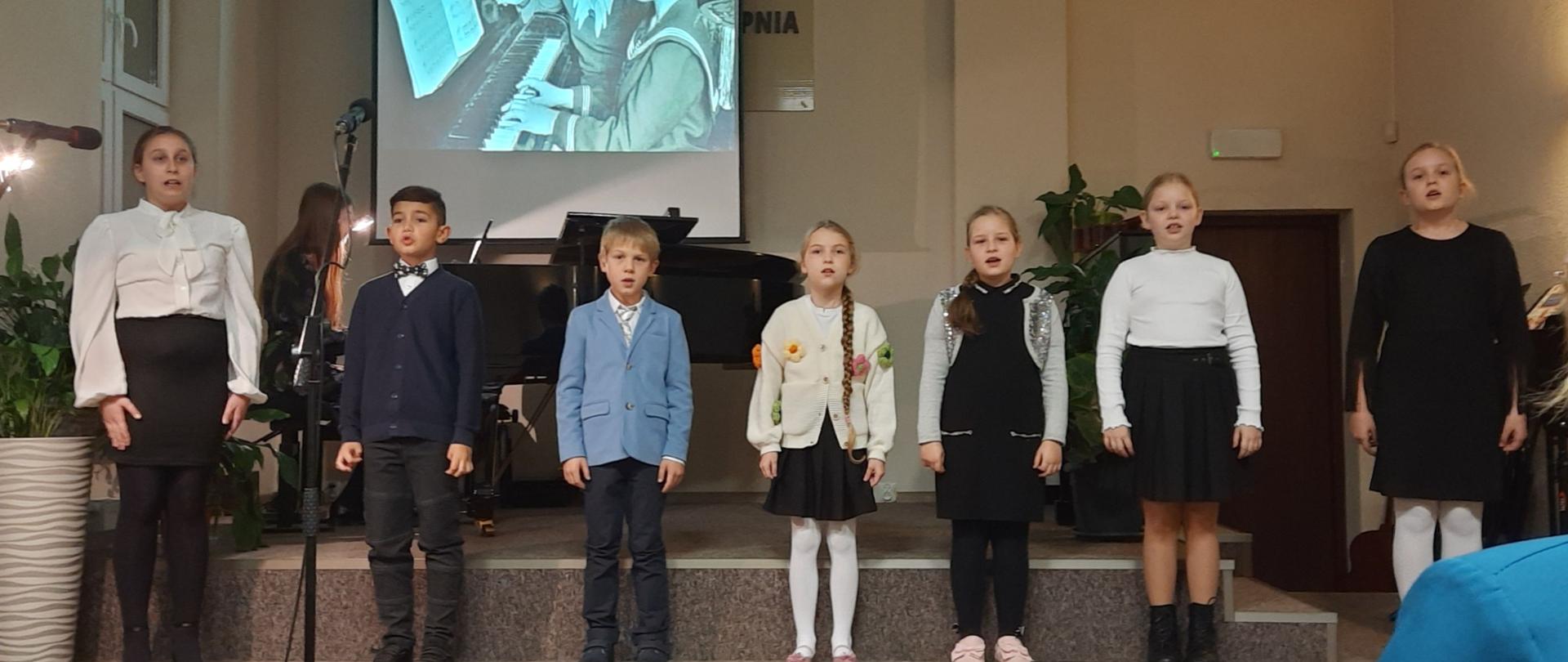 Dzieci z chóru szkolnego stoją na scenie w śpiewają piosenkę