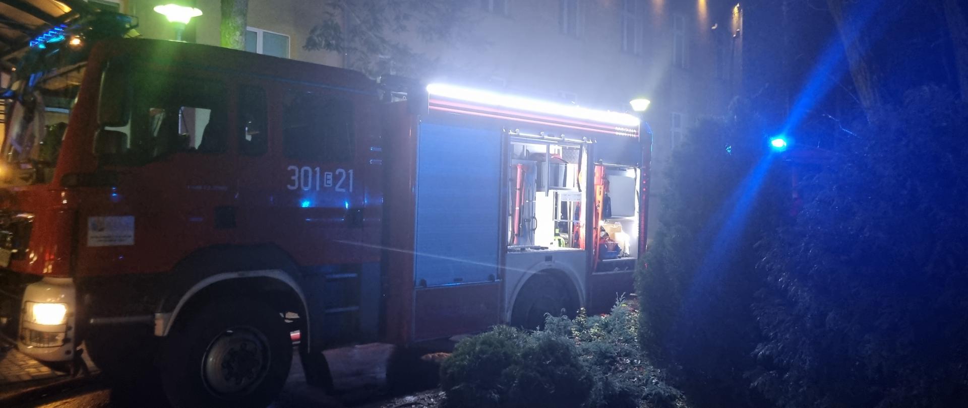 Samochód gaśniczy z otwartymi skrytkami bocznymi stojący przed budynkiem szpitala