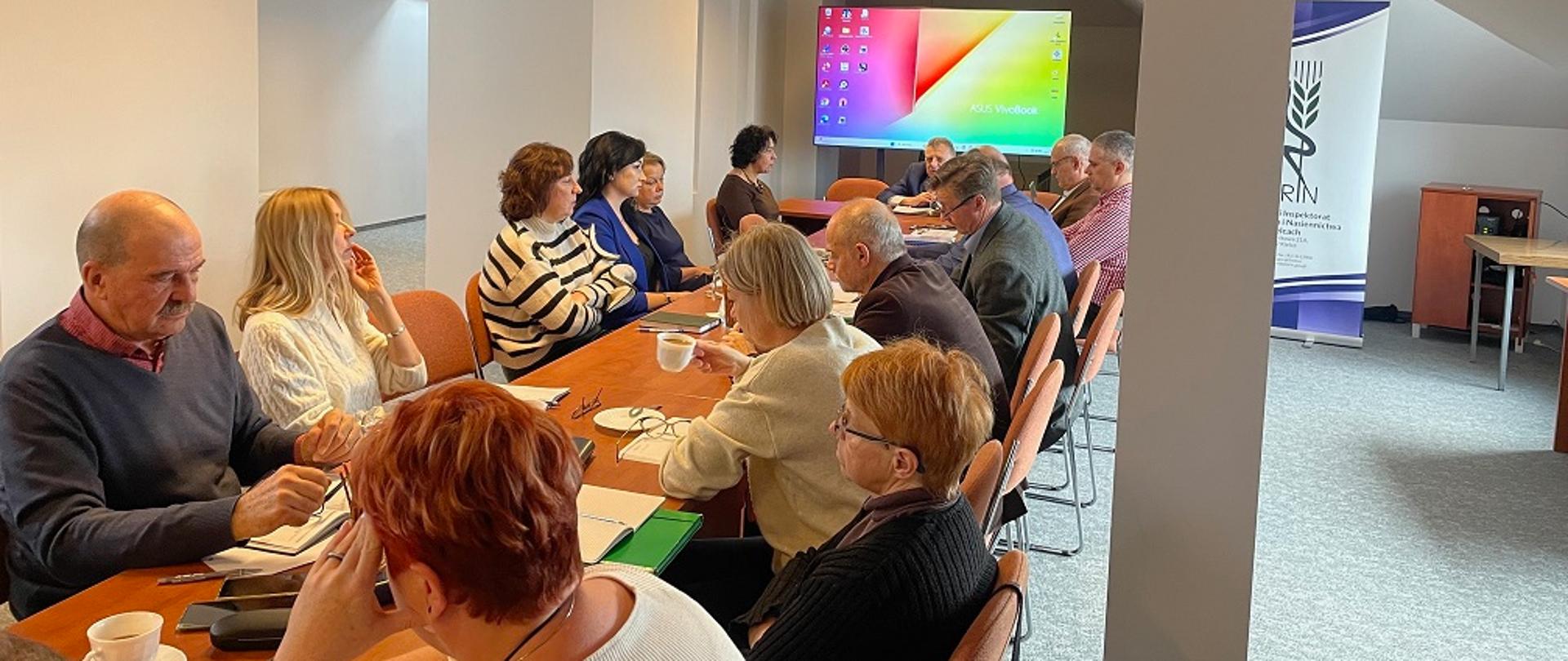 Przy podłużnym, rudym stole narad widać 15 osób usadzonych po prawej i po lewej stronie- mężczyźni i kobiety. U szczytu siedzi przemawiający mężczyzna. Każda z osób jest zamyślona, zasłuchana w słowa mówcy. Na stole znajdują się notatniki, długopisy i filiżanki z kawą. Za głową przemawiającego widoczny jest rzutnik . Po prawej stronie zdjęcia stoi roll-up z logo Państwowej Inspekcji Ochrony Roślin i Nasiennictwa. Cała sceneria ukazana jest w jasno szarej barwie ścian sali konferencyjnej. 