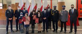 Na zdjęciu czworo młodych bohaterów z otrzymanymi medalami. Na zdjęciu z młodymi bohaterami znajdują się Wicewojewoda Kujawsko-Pomorski, Kujawsko-Pomorski Komendant Wojewódzki PSP, Zastępca Komendanta Wojewódzkiego Policji w Bydgoszczy, Dyrektor Wydziału Bezpieczeństwa i Zarzadzania Kryzysowego KPUW, Prezes Nadgoplańskiego WOPR, Burmistrz Kruszwicy, Zastępca Prezydenta Torunia oraz Zastępca Burmistrza Koronowa. Zdjęcie wykonano w garażu na tle flag oraz samochodu ratowniczo-gaśniczego.