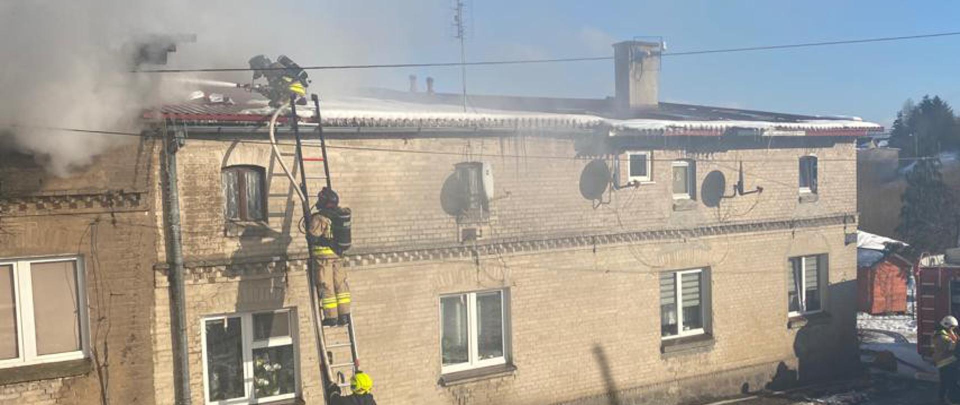Widok domu od ulicy. Spod dachu wydobywa się dym. Strażacy podają prąd wody z drabiny, z prawej strony widać strażaka obsługującego autopompę samochodu pożarniczego.