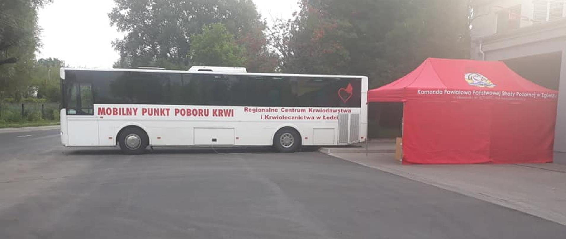 Zdjęcie mobilnego punktu poboru krwi, stacjonującego podczas akcji krwiodawstwa przed Komendą Powiatową PSP w Zgierzu, obok autokaru z Regionalnego Centrum Krwiodawstwa i Krwiolecznictwa w Łodzi znajduje się czerwony namiot Komendy Powiatowej Państwowej Straży Pożarnej w którym odbywa się wstępna kwalifikacja i podpisanie deklaracji osób oddających krew.